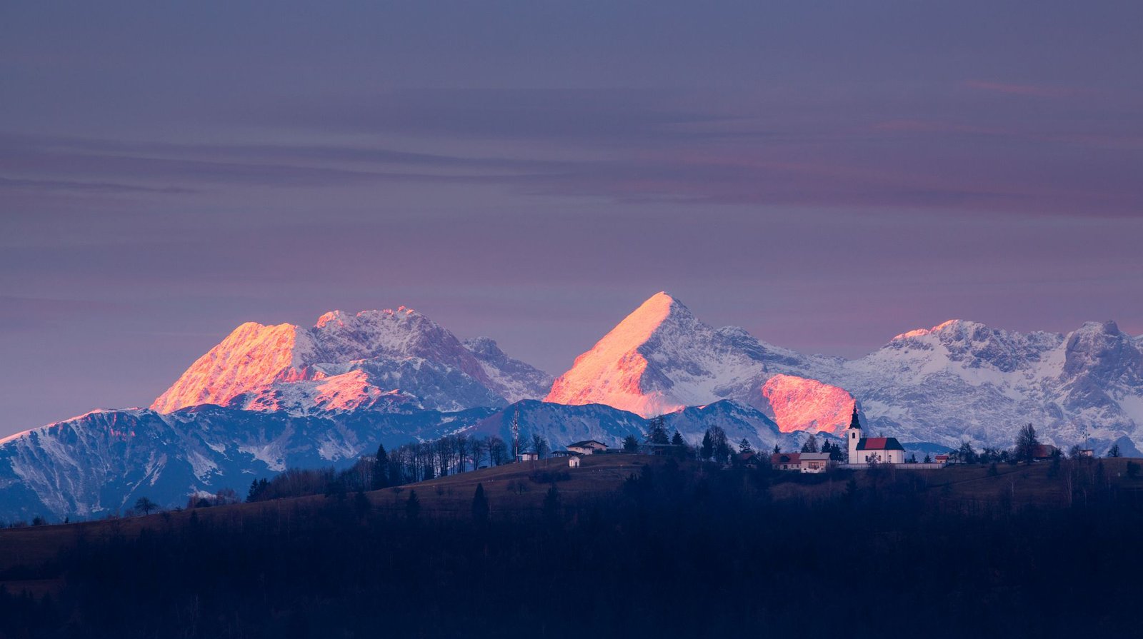 View across to the church of Saint Nicholas and the village of Jance at sunset with the Kamnik Alps in the background, in the hilly region to the east of Ljubljana, Slovenia. Seen from Mali Vrh village.
